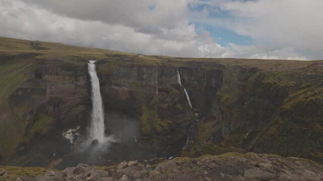 Haifoss plunges into a deep rugged canyon beside a slender neighboring waterfall under broken clouds, establishing overview near edge