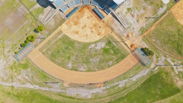 : A drone shot looks straight down on a baseball diamond, showing the sandy infield, curved outfield boundary, and surrounding green grass. Covered stadium seating with blue bleachers wraps.
