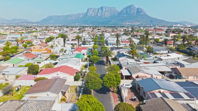 A drone shot looks down a straight, palm-lined suburban street on the Cape Flats, with rows of single-story homes featuring colorful facades and red-tiled roofs stretching toward the iconic silhouette