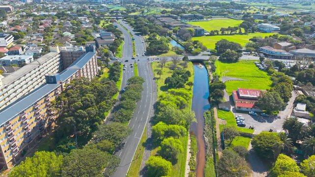 A drone shot captures a curving suburban road lined with trees, running parallel to a river canal that flows past apartment blocks, sports fields, and a red-roofed commercial building with a parking