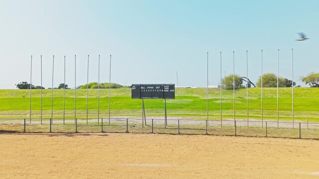 A drone shot swoops in toward a manual baseball scoreboard displaying "At Bat," "Ball," "Strike," "Out," "Pitch Timer," "Runs," and "Hits" fields, positioned on a wide, open grass field flanked by row