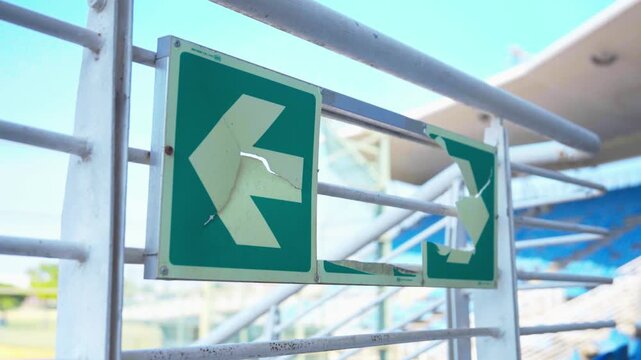 : A close-up shot captures two weathered, cracked green directional arrow signs mounted on metal railings within a stadium, with faded paint and visible wear suggesting years of exposure.