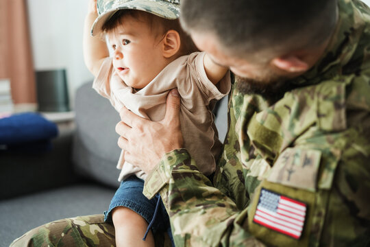 Male american soldier in military uniform hugs his little son at home while baby boy playing with his hat - Emotional happy homecoming concept - Main focus on man hand