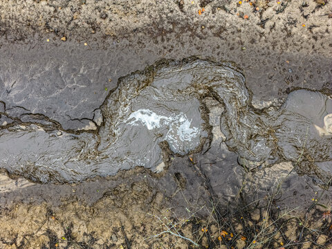 Aerial view of a small, muddy stream flowing through wet, textured soil with swirling water patterns and patches of dry earth in Latvia.