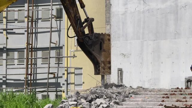 Close-up shot of a heavy excavator equipped with a hydraulic breaker attachment actively breaking down concrete structures and debris at a construction or demolition site during the daytime. 