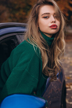 Young woman with red lipstick in green sweater leaning on a blue car outdoors