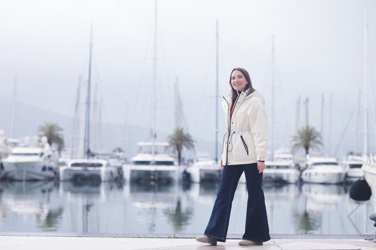 Woman in White Coat Posing at Marina with Yachts and Sailboats on a Cloudy Day