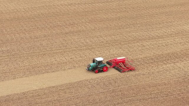 Crop planting tractor with red seeding implement moving through harvested farmland. Parallel stubble rows across extensive agricultural field. Mechanized sowing equipment for efficient grain
