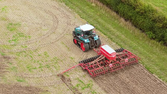 Agricultural seeder working close to grassy field margin and woodland hedge. Tracked tractor pulling wide planting implement across stubble. Modern crop cultivation on arable farmland beside green