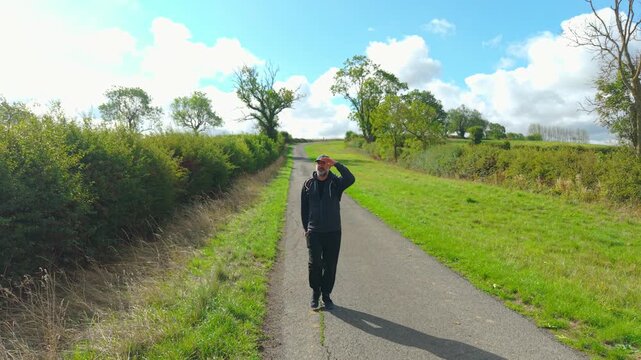 Countryside traveler with backpack walking along winding rural road under bright blue sky. Middle aged man hiking between green hedgerows and open fields. Outdoor fitness and active lifestyle during