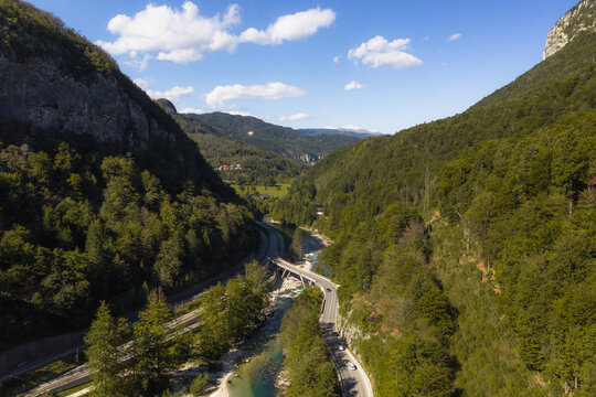 Aerial view of Sava Bohinjka river flowing through a lush green valley with a road bridge and forested mountain slopes under a clear blue sky, Radovljica, Slovenia.