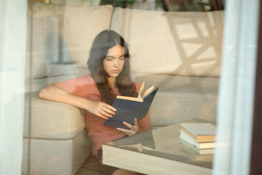 Young woman reading paper book viewed through a window