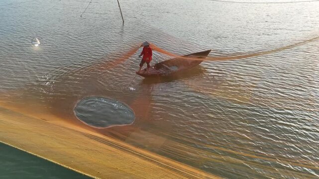 Aerial view of a fisherman in a small wooden boat casting a large fishing net on the Thu Bon River in Hoi An, Da Nang, Vietnam.