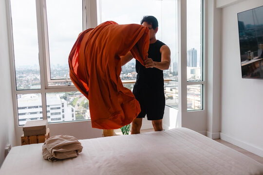 Man making bed in modern apartment with city view, spreading orange sheet
