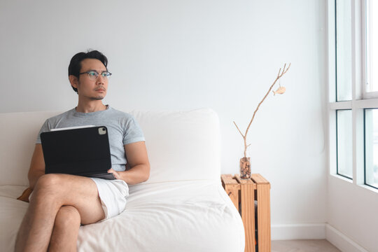 Thoughtful Asian Man in Glasses Sitting on Sofa with Tablet at Home
