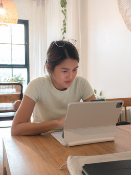 Focused Young Asian Woman Working on Her Tablet with a Keyboard at a Wooden Table.