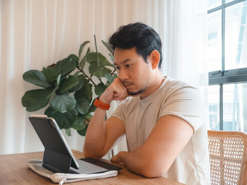 Pensive Asian Man Concentrating on His Digital Tablet While Working Remotely from Home