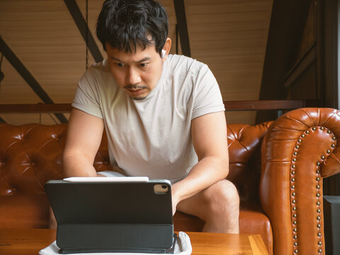 Focused Asian man intently uses digital tablet with stylus on a leather sofa