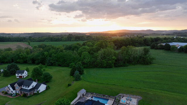 Aerial view of a residential property with a swimming pool and a large house surrounded by lush green fields and trees at sunset in Elizabeth Township, Pennsylvania, United States.