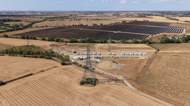 Aerial view of the Eaton Socon Substation and adjacent solar farm with rows of photovoltaic panels and a transmission tower in the rural landscape of St. Neots, England, United Kingdom.