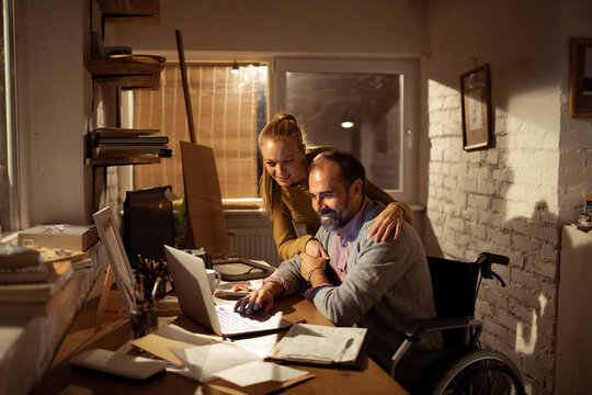 Woman hugs wheelchair user while working on laptop at home