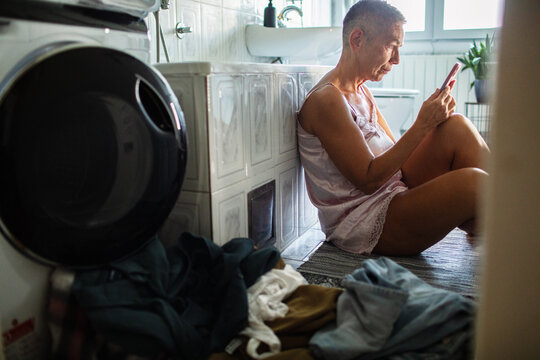 Woman sitting on bathroom floor texting by washing machine
