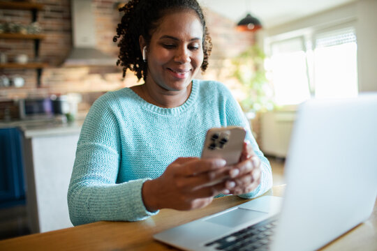 Smiling woman using smartphone with earbuds at kitchen table