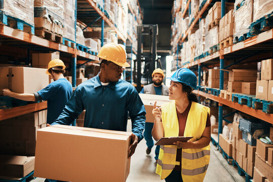 Worker with clipboard guides coworker carrying box in warehouse