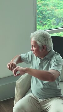 50s middle aged southeast asian man sitting in armchair, stretching arms to stay awake in living room, vertical video