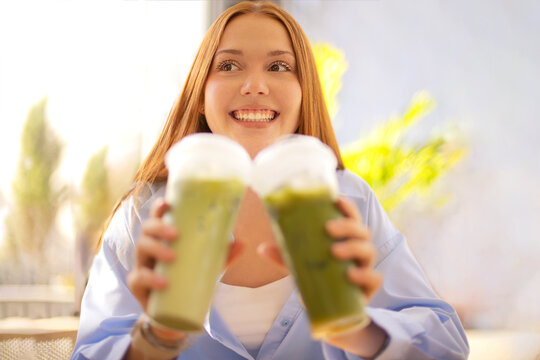 Smiling woman holding two green smoothies toward camera with bright fruit decor and softly blurred indoor background, fresh beverage portrait in a sunlit interior setting