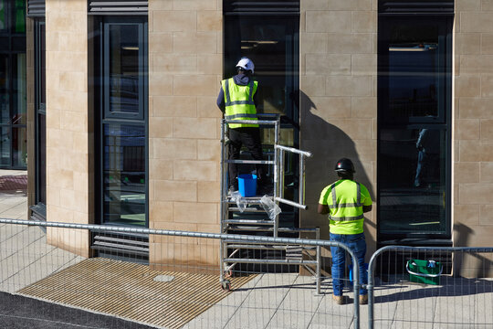 Window cleaning worker performing commercial building facade maintenance from a mobile scaffold tower. The elevated work area includes safety equipment, protective clothing, cleaning supplies