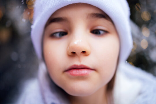 Young girl with light brown hair wearing a white hat and coat, close-up portrait with snowflakes falling in a winter setting, soft bokeh background