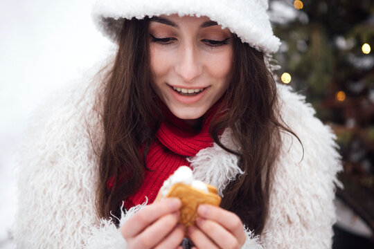 Young woman with long brown hair wearing a white fur coat and hat holds a pastry while smiling outdoors in a winter setting with blurred lights in the background