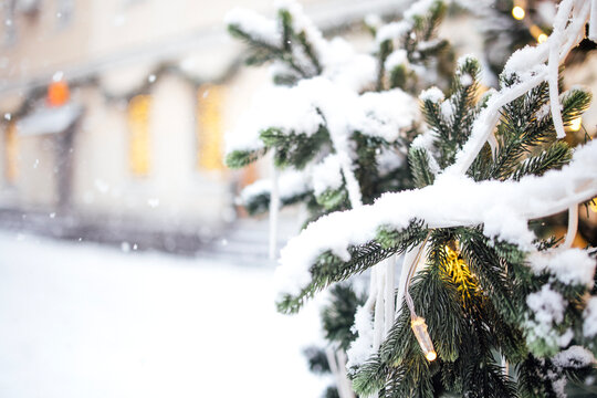 Snow-covered Christmas tree adorned with white ribbons and warm lights, set against a softly blurred winter street scene with festive decorations