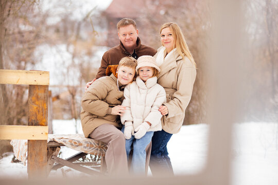 Family of four posing together outdoors in winter clothing, seated on a wooden bench in a snowy landscape with trees and a house in the background