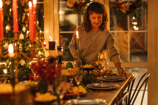 Woman setting a festive dining table with plates and decorations for a holiday meal, Christmas tree and candles visible in a cozy indoor setting