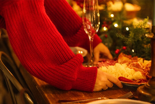 Hands of a person in a red sweater arranging a festive charcuterie board with assorted meats and cheeses on a wooden table with holiday decorations and glassware
