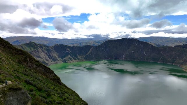 Aerial view of the turquoise Quilotoa Lake crater surrounded by steep volcanic walls and lush green slopes under a cloudy sky in Cotopaxi, Ecuador.