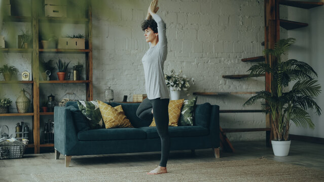 Flexible and strong young woman is training at home doing yoga standing on one leg maintaining balance with raised arms. Healthy lifestyle, house and interiors concept.