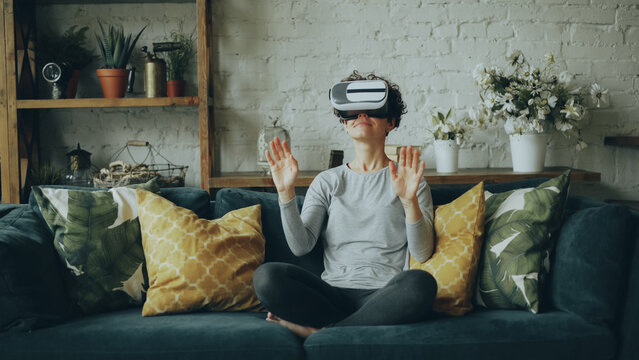 Zoom-in of smiling young lady in augmented reality glasses moving hands and turning head sitting on sofa at home and having fun. Entertainment and modern technology concept.
