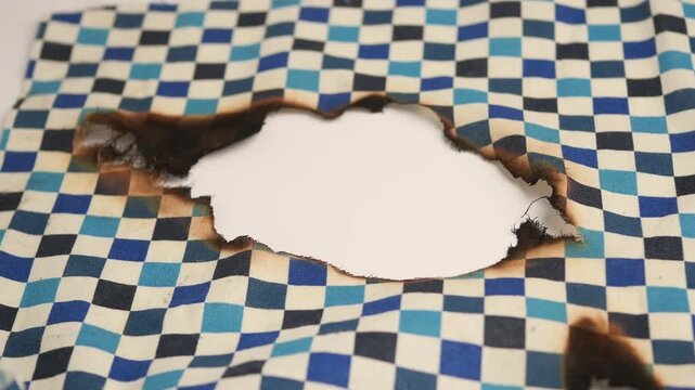 Burned hole in blue and white checkered fabric background close up footage.