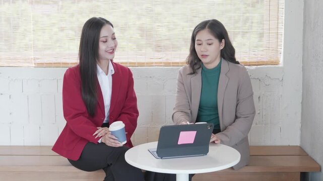 Two Asian businesswomen sitting at a small office table, discussing work details using a digital tablet and taking notes on paper documents during a business meeting near a large window.
