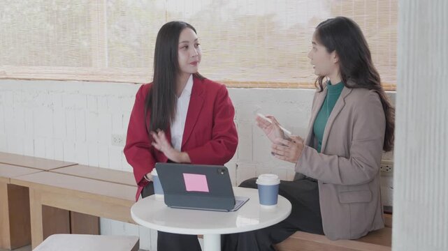 Two Asian businesswomen sitting at a small office table, discussing work details using a digital tablet and taking notes on paper documents during a business meeting near a large window.