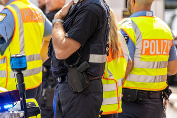 German police officers in high-visibility yellow vests stand outside next to a patrol car during an operation. In the background, more police vehicles are visible on the street during daytime. © Thomas