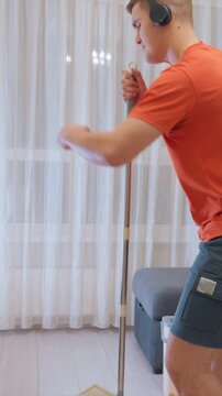 Man with black socks and blue shorts uses flat mop to clean smooth white wooden flooring. Man moves back and forth across different sections of floor while wearing headphones