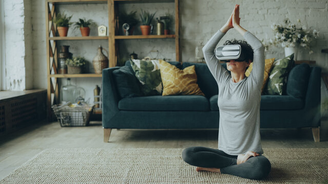 Slim young lady in artificial reality glasses is meditating at home in lotus pose with hands in namaste enjoying simulation and relaxing. Modern technology, people and experience concept.
