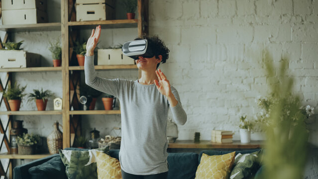 Happy girl is using virtual reality glasses at home standing in light room moving hands and smiling having fun. Modern gadgets, entertainment and apartment concept.