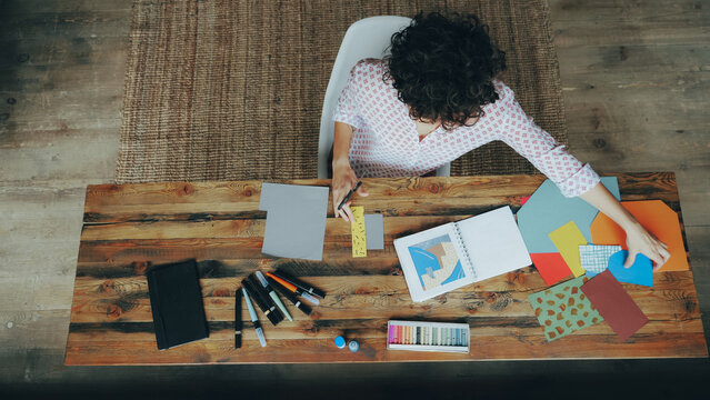 Top view of slim curly-headed woman making colorful paper collage sitting at table cutting figures with scissors. Design, creative people and interiors concept.