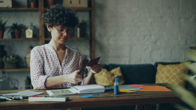 Creative young woman is cutting piece of paper with scissors creating collage then putting it in notebook and checking design. Creativity, workspace and modern art concept.