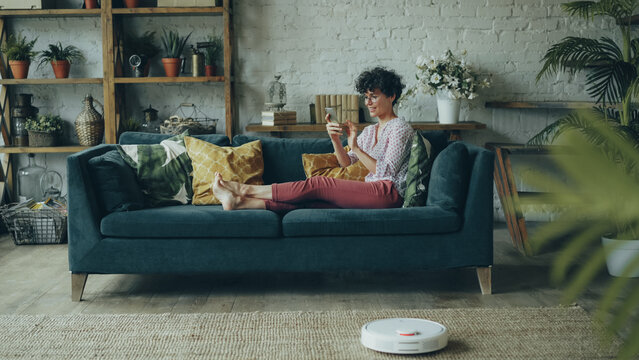 Happy relaxed girl is holding smartphone touching screen and smiling resting on couch while robotic vacuum cleaner is doing housework cleaning apartment floor.